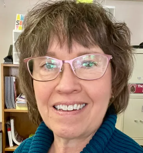 joanie.png A woman with glasses smiling indoors, with books and shelves in the background.