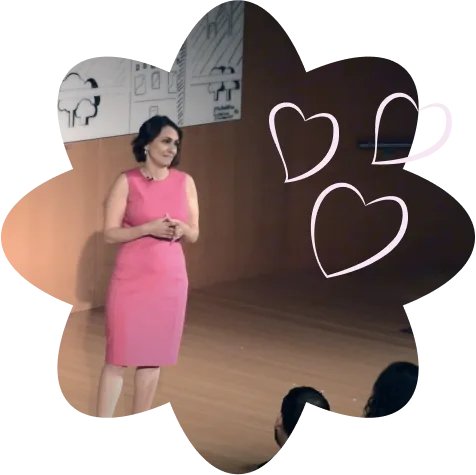 Group+12-2.png Laurie Gerber speaks on a stage in a pink dress, framed by a flower shape with heart line drawings.