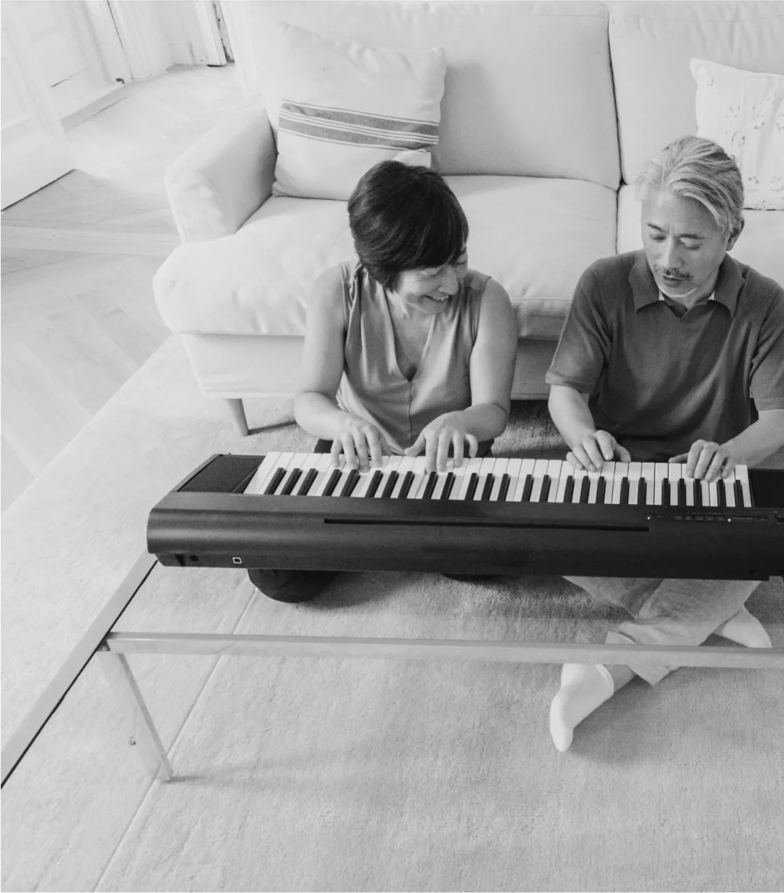 An older couple sit on the floor playing an electric keyboard together in a bright living room, in black and white.